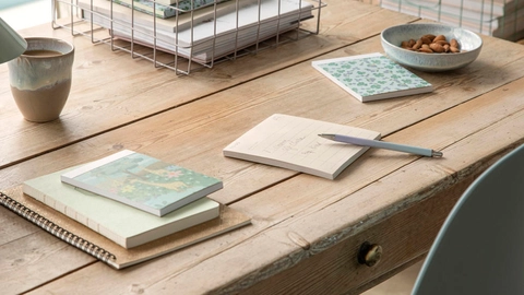 Et træbord med en kop kaffe, notesbøger i forskellige størrelser, en kuglepen, og en skål med mandler. I baggrunden er der en metaltrådskurv med bøger.